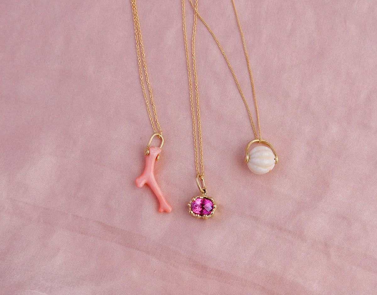 Three pendants including a natural pink coral branch, a bright pink tourmaline gemstone and a hand-carved vintage coral bead on 14kt gold bails lay on pink silk background.