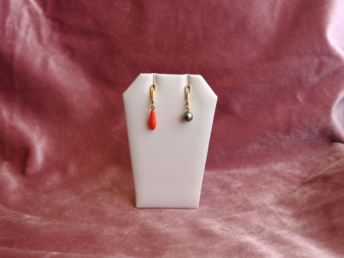 A pair of small gold drop earrings displayed on a white jewelry stand against a dusty rose velvet fabric background. One earring has a bright coral-red teardrop bead, and the other has a small dark gray pearl.