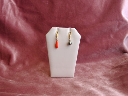 A pair of small gold drop earrings displayed on a white jewelry stand against a dusty rose velvet fabric background. One earring has a bright coral-red teardrop bead, and the other has a small dark gray pearl.