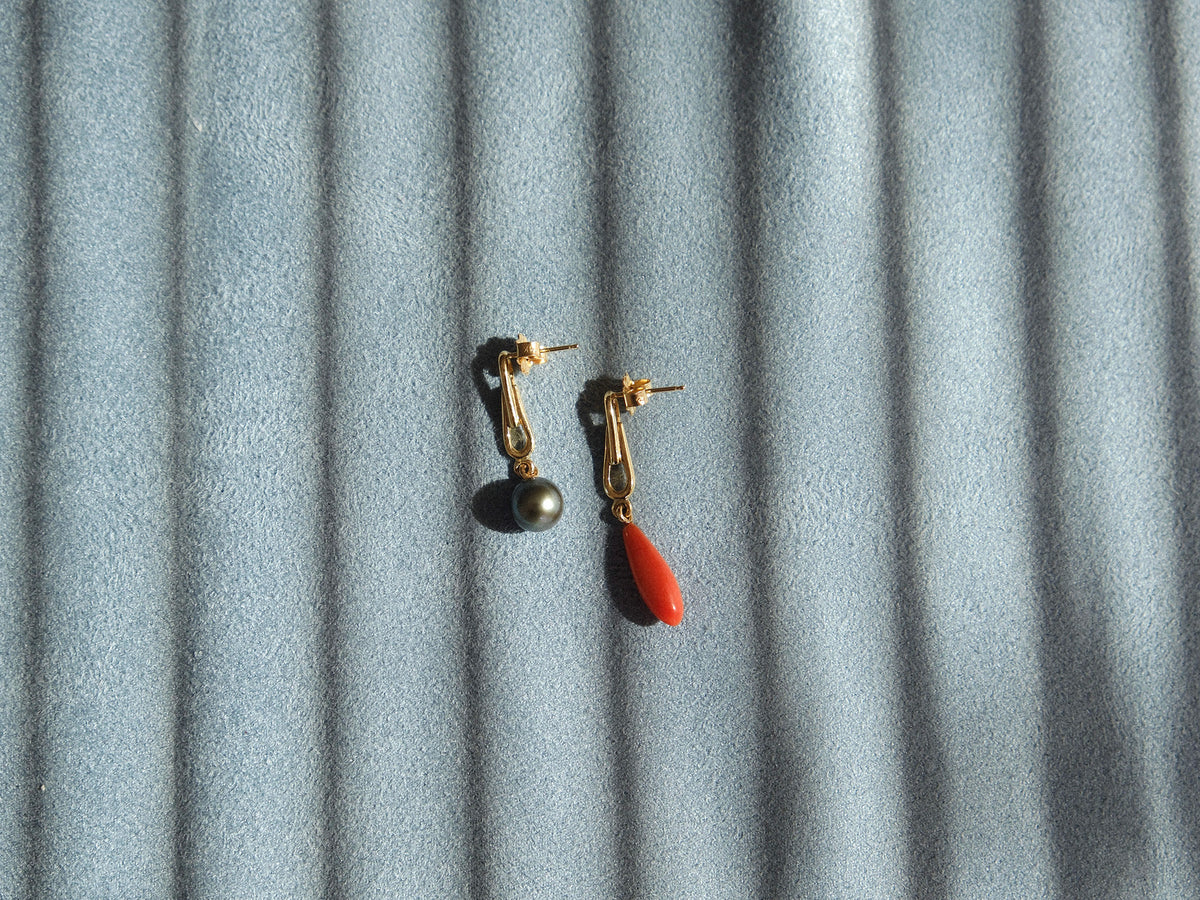 A pair of gold dangle earrings, one with a dark gray round pearl and the other with a small orange-red teardrop coral, displayed on a textured gray fabric background with diagonal shadows.