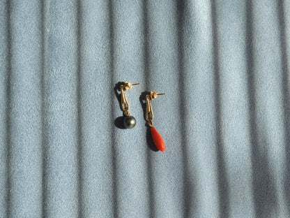A pair of gold dangle earrings, one with a dark gray round pearl and the other with a small orange-red teardrop coral, displayed on a textured gray fabric background with diagonal shadows.