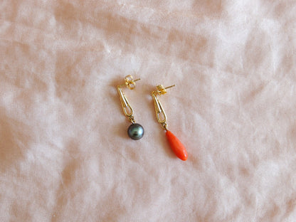 A pair of gold drop earrings on pink silk. One earring features a small dark gray pearl, and the other features a bright coral drop.