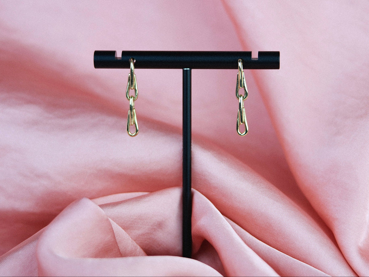 Gold chain earrings on a black stand against a pink fabric background