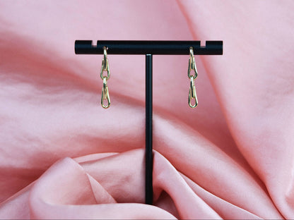 Gold chain earrings on a black stand against a pink fabric background