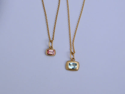 Two gold chain necklaces with rectangular gemstone pendants, one featuring a pink stone and the other a light blue stone, displayed against a light lavender background.