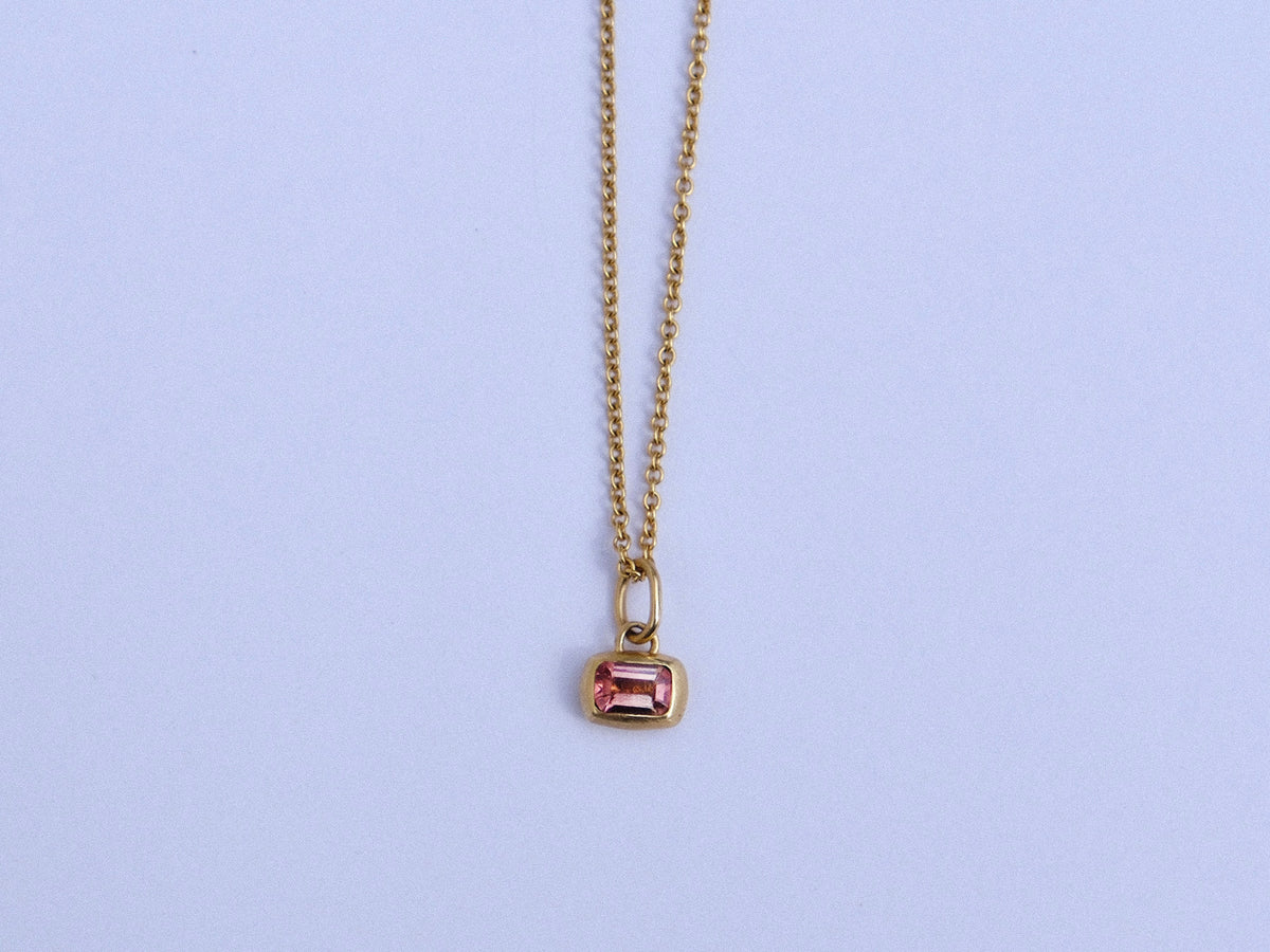 A delicate gold chain necklace with a small rectangular gold pendant featuring a pink gemstone, displayed against a light background.