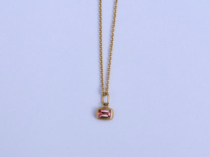 A delicate gold chain necklace with a small rectangular gold pendant featuring a pink gemstone, displayed against a light background.