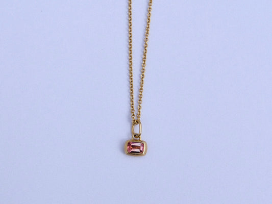 A delicate gold chain necklace with a small rectangular gold pendant featuring a pink gemstone, displayed against a light background.