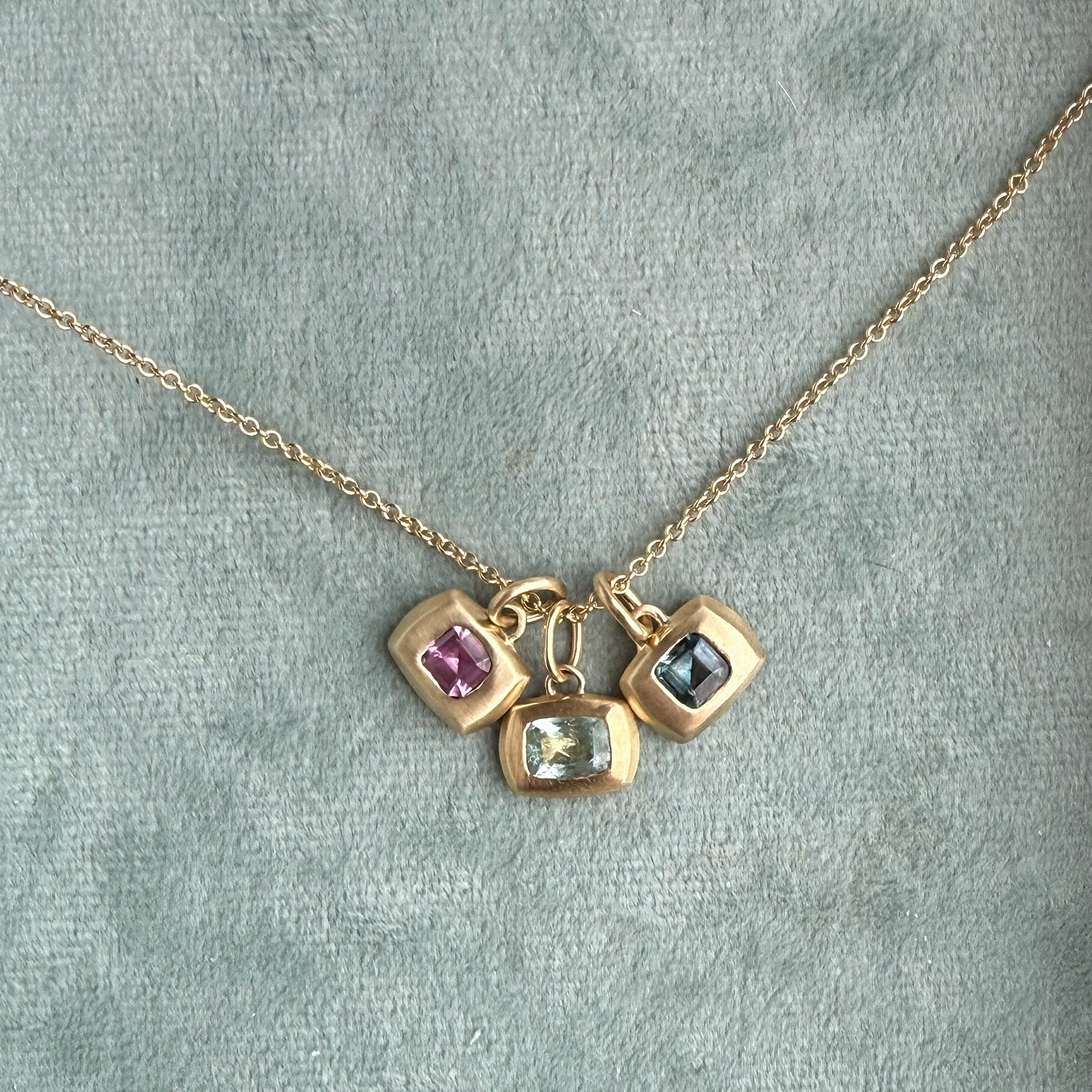 A gold chain necklace with three small square charms set with different colored stones, displayed on a textured gray background.