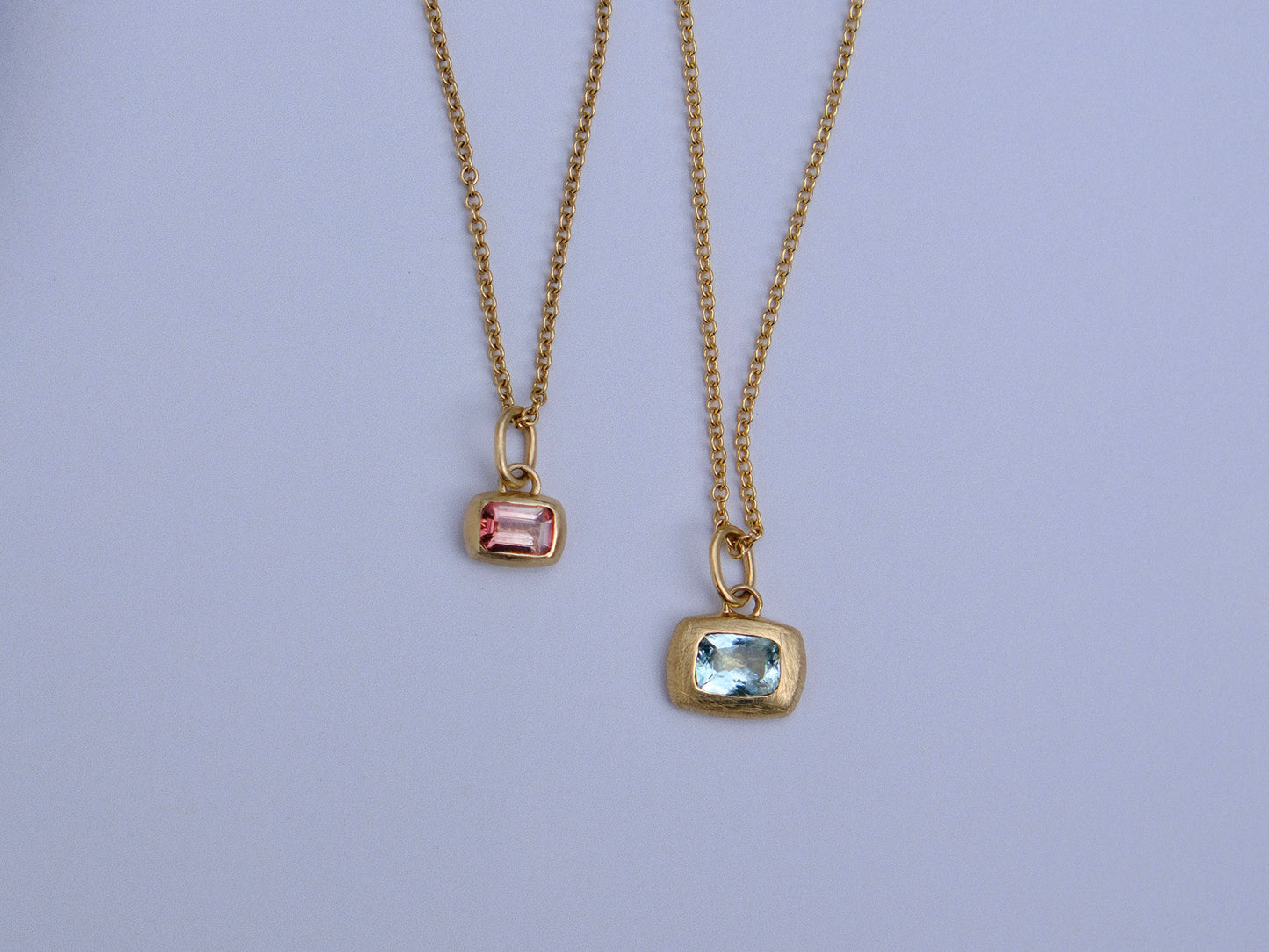 Two gold chain necklaces with rectangular gemstone pendants, one featuring a pink stone and the other a light blue stone, displayed against a light lavender background.