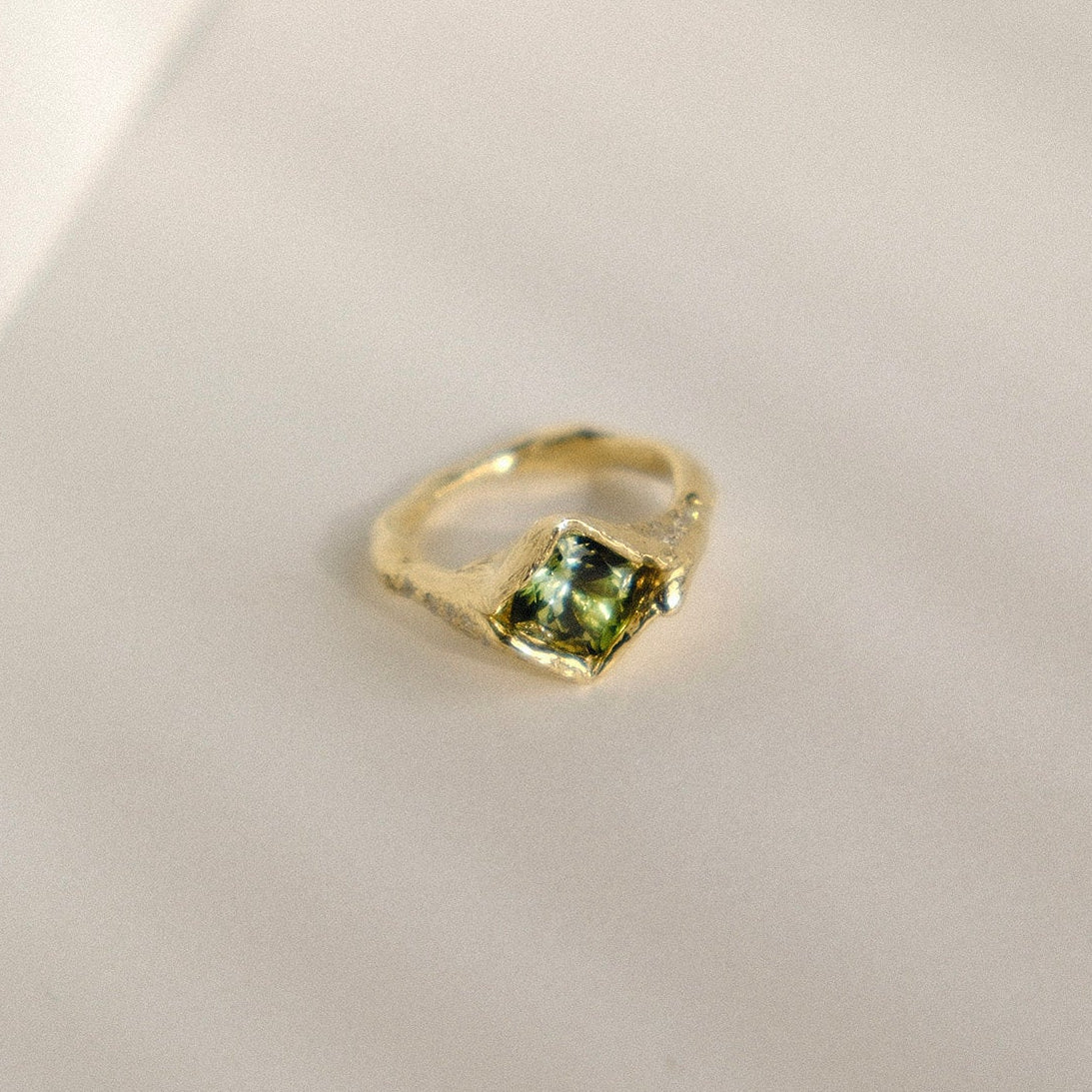 A gold ring with a square-cut green gemstone, resting on a light beige textured surface.