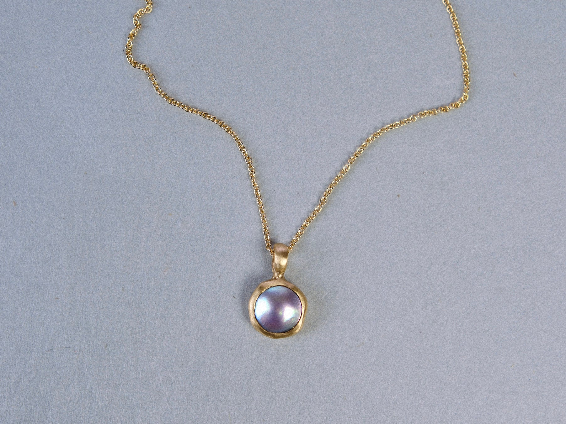 A gold chain necklace with a round bezel-set pendant featuring a lustrous lavender-grey pearl, displayed on a neutral gray background.