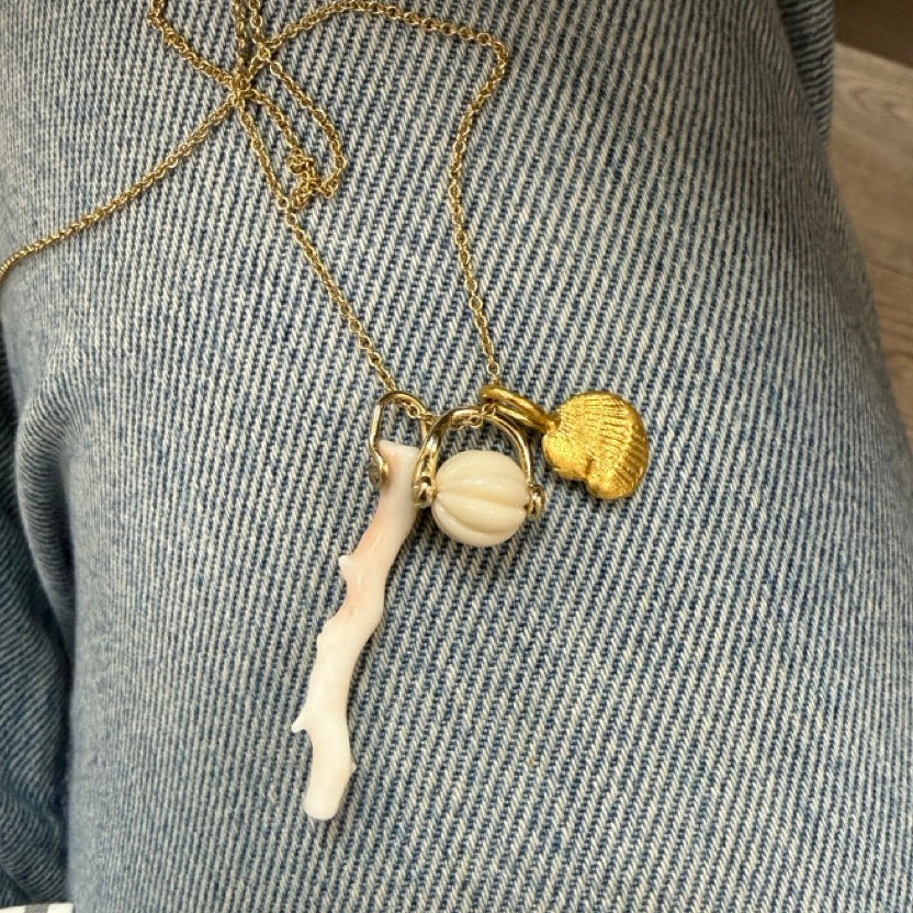 A close-up of a gold chain necklace with a white shell pendant, light pink coral branch pendant, and a small gold seashell charm, resting on blue denim jeans.