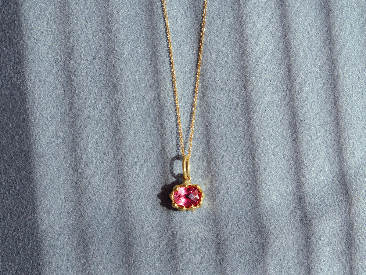 A gold chain necklace with a small oval pink tourmaline pendant, displayed against a textured blue background with soft diagonal shadows.