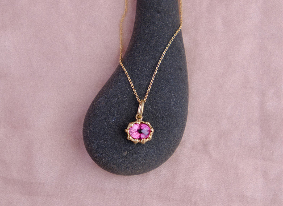 A gold chain necklace with an oval pink tourmaline pendant, displayed on a dark gray stone against a soft pink background.