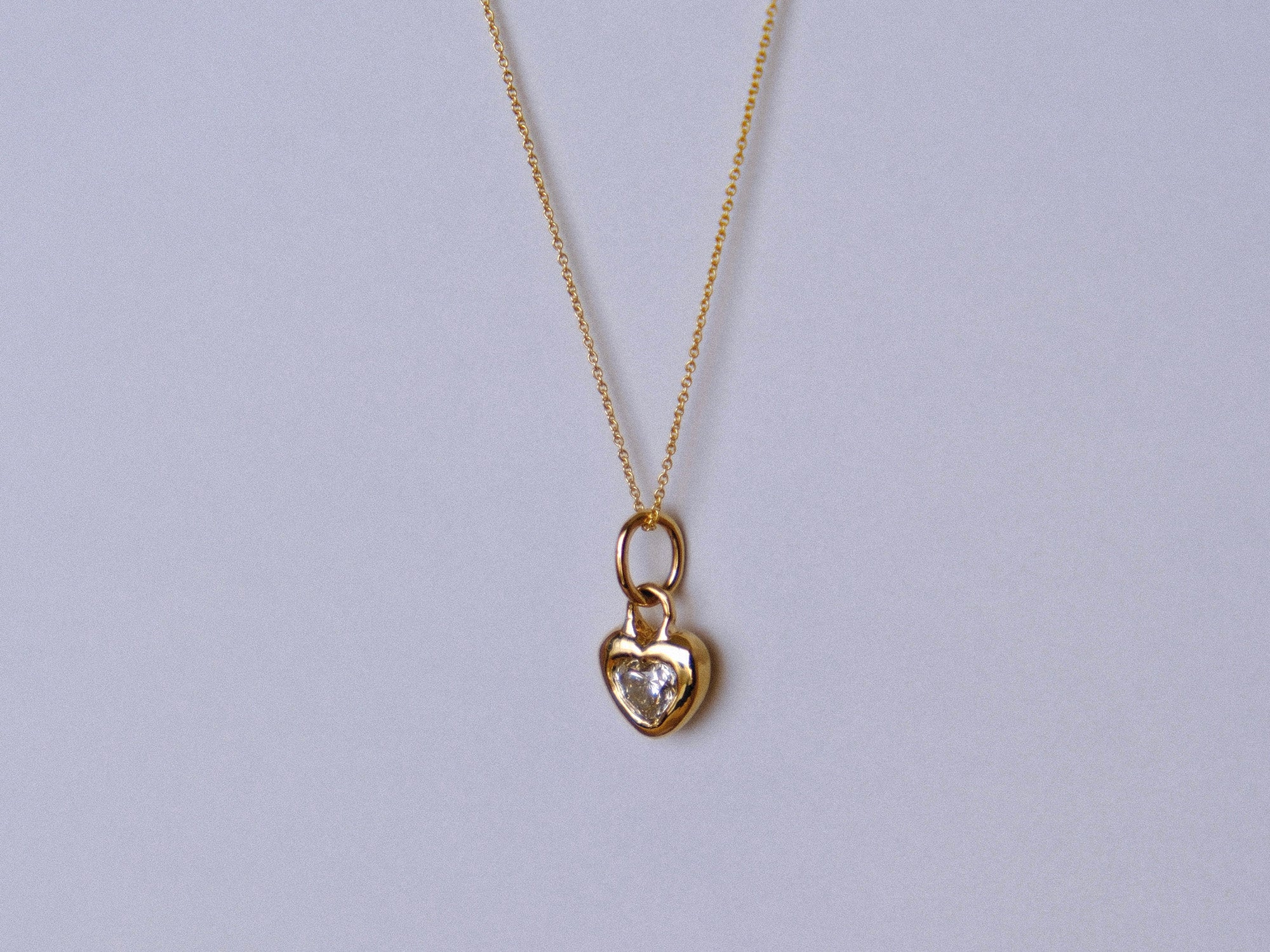 A delicate gold chain necklace with a small heart-shaped pendant featuring a clear heart-shaped gemstone, displayed against a light gray background.