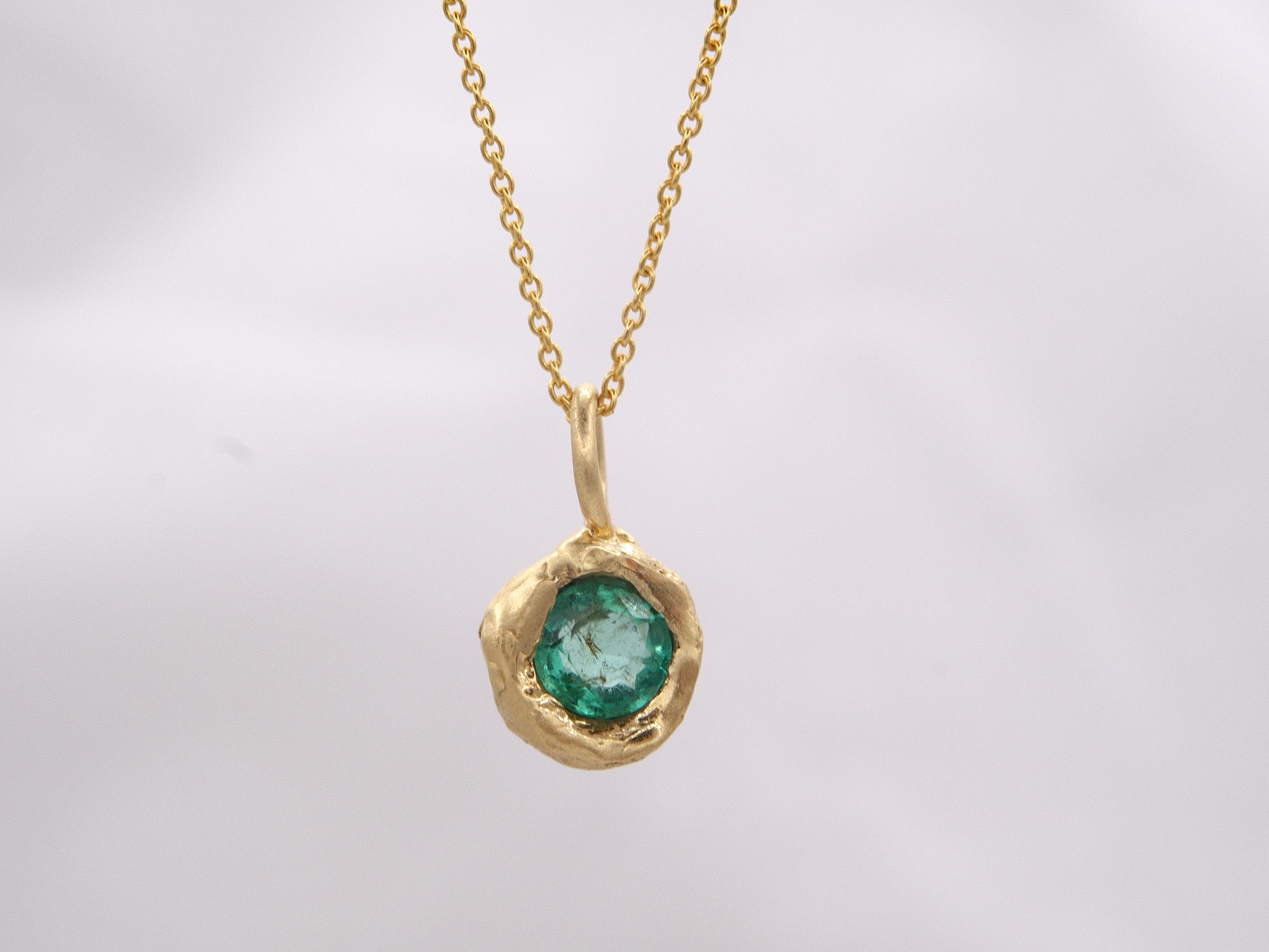A gold pendant with an emerald embedded in a gold setting, featuring an organic texture on a chain.