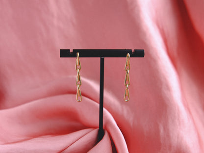 Gold chain earrings dangle on a black earring stand against a pink fabric background