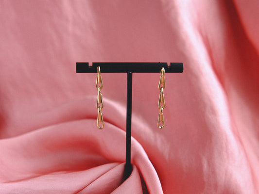 Gold chain earrings dangle on a black earring stand against a pink fabric background