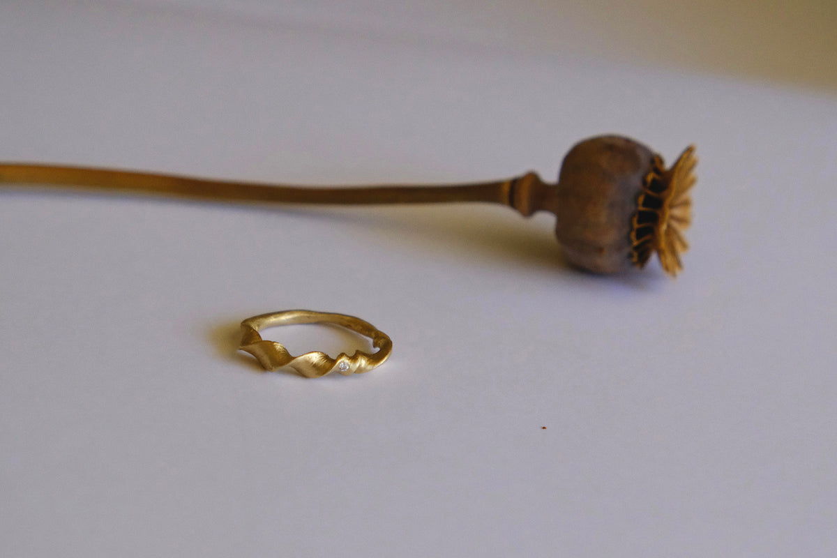 Organic twist ring with a single white diamond laying on a gray background next to a poppy.
